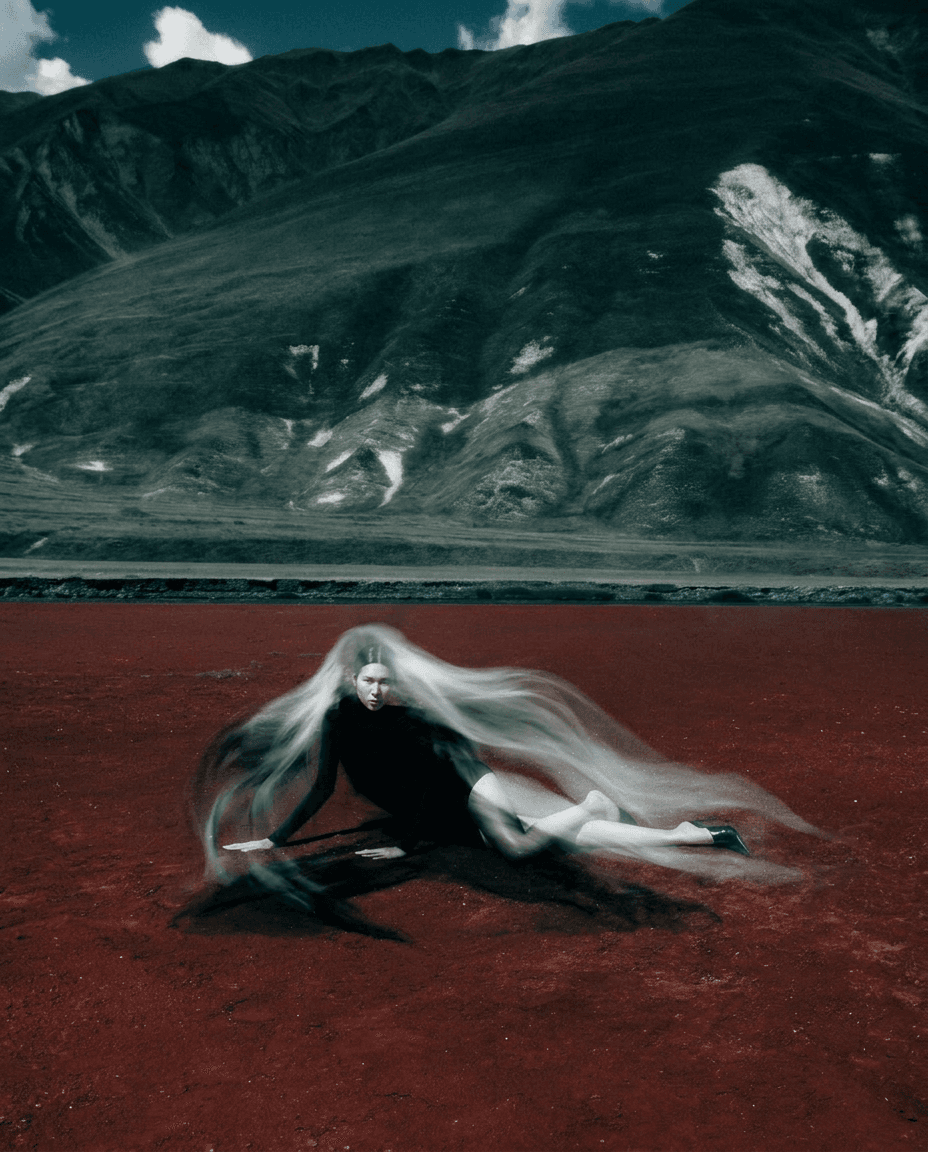 A woman with long hair poses on a red landscape with mountains behind her.