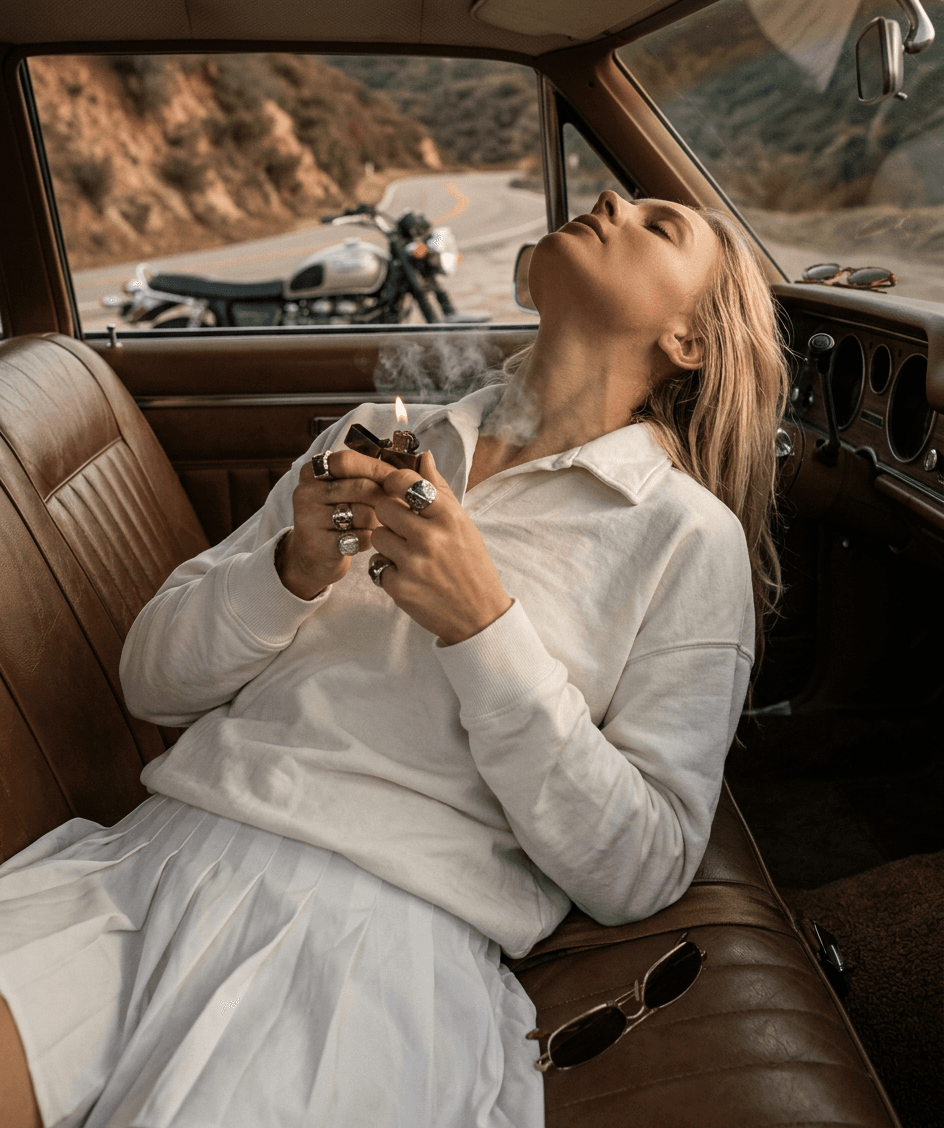 Woman reclining in a classic car, holding a lighter and looking up.