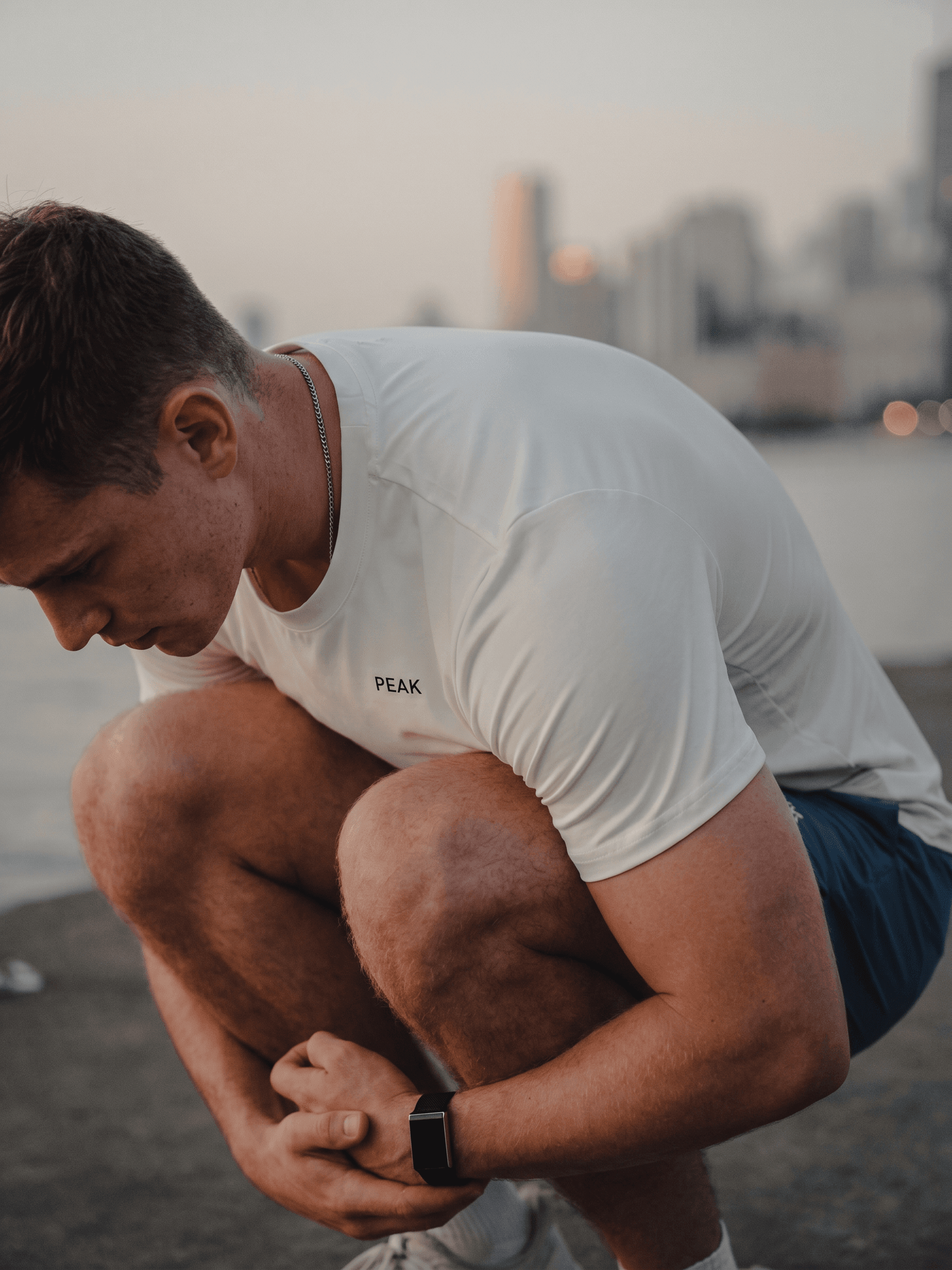 A man squatting near a waterfront at sunset with blurred city buildings.