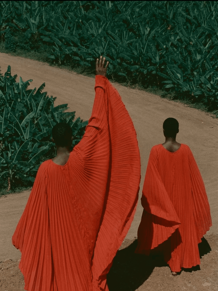 Two figures in red clothing walking on a dirt path with greenery around.