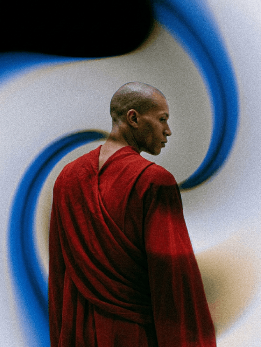 A man wearing a red robe with a surreal blue and beige swirled background.