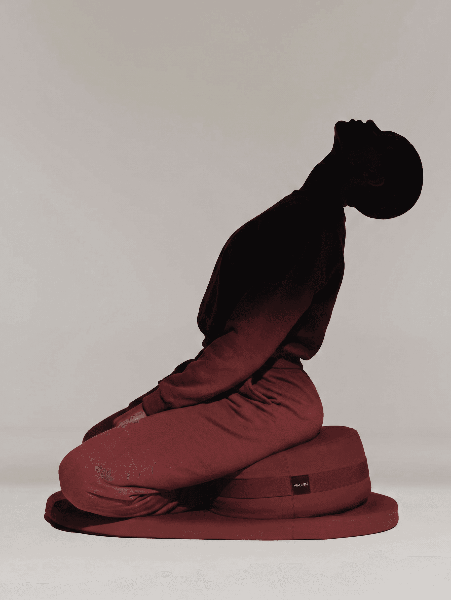 Silhouette of a woman in a red outfit kneeling on a cushion with a grey background.