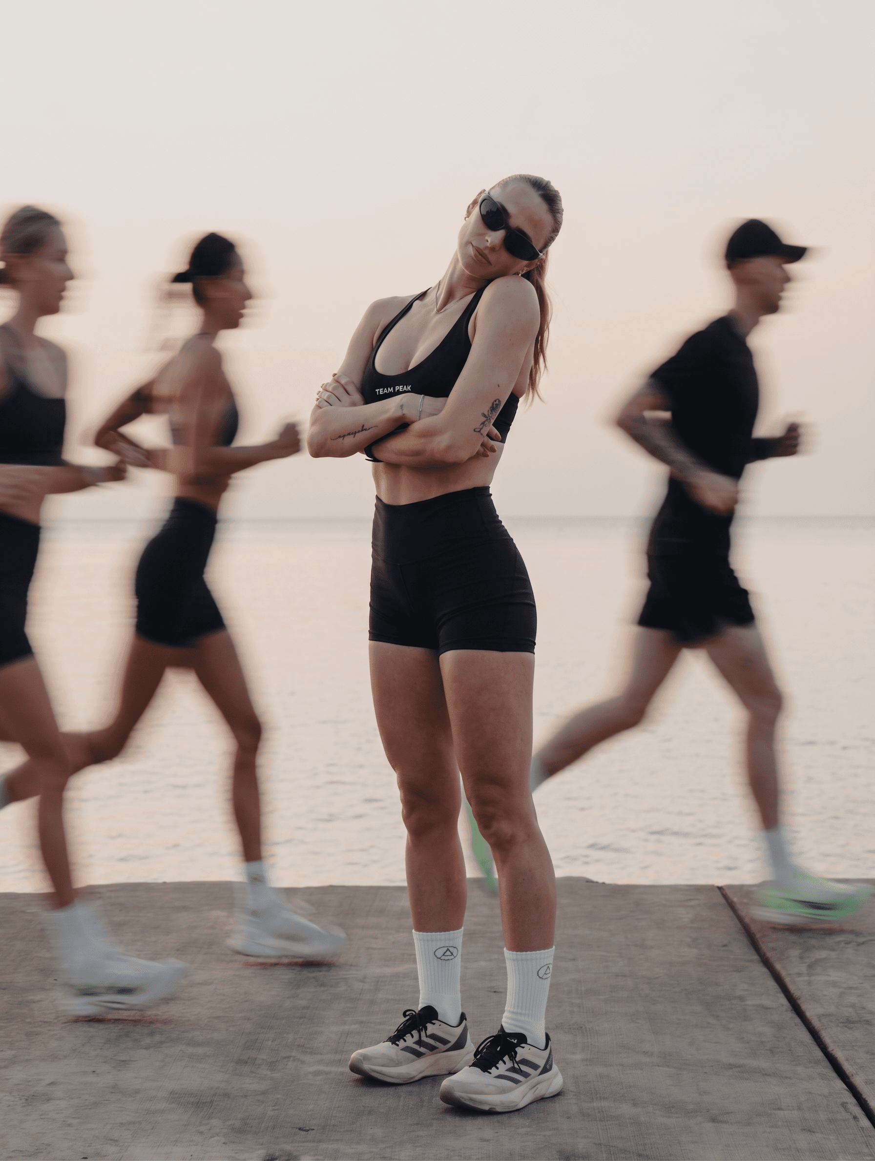 A confident woman stands in front of a calm water body with blurred runners in the background.