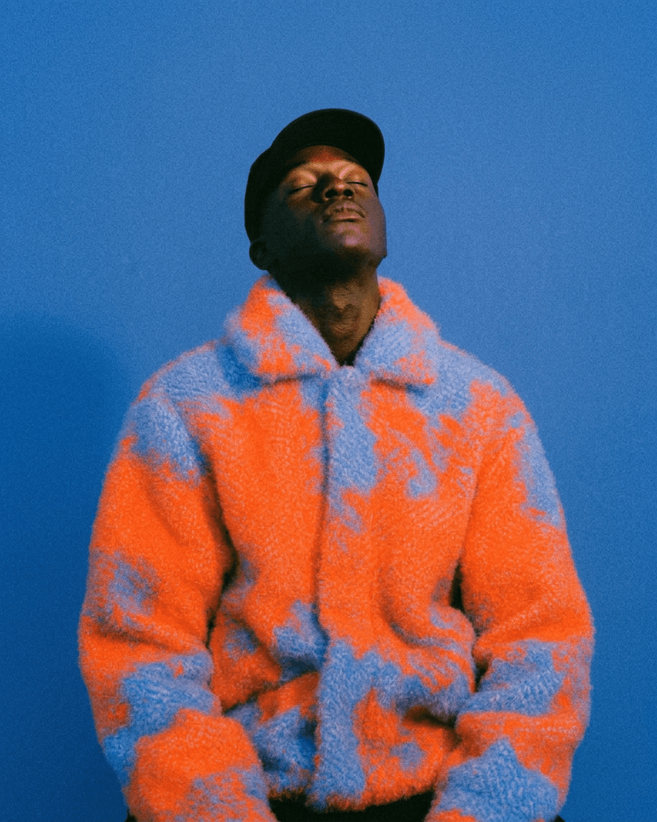 A man wearing a vibrant oversized fuzzy jacket stands against a bright blue background.