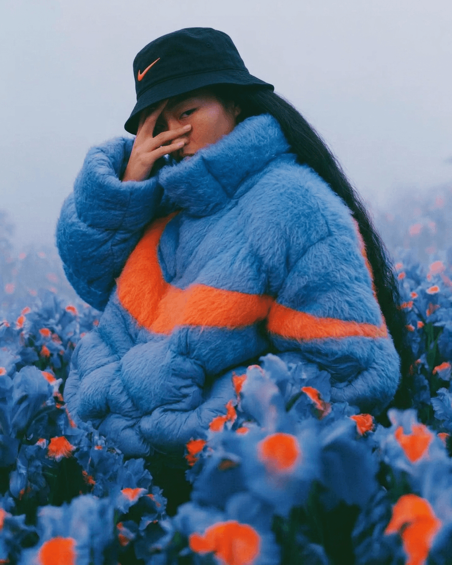 Woman in a blue jacket and bucket hat poses among blue and orange flowers.