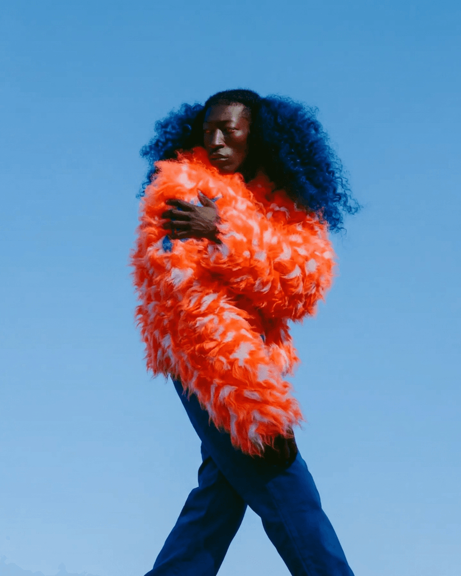 Full-figure portrait of a model wearing an orange feathered coat with a blue sky background.