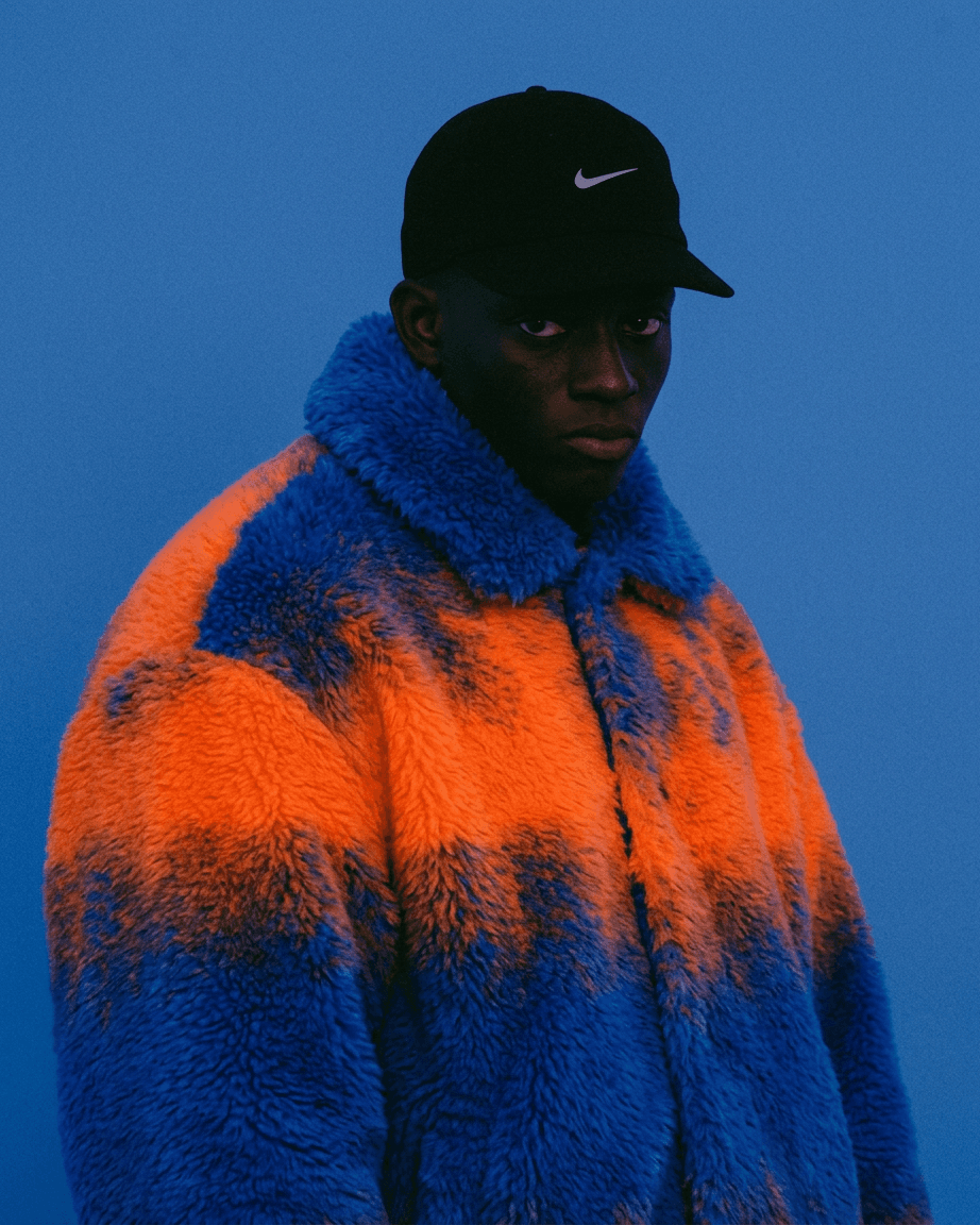 Portrait of a man wearing a fluffy orange and blue jacket with a blue background.