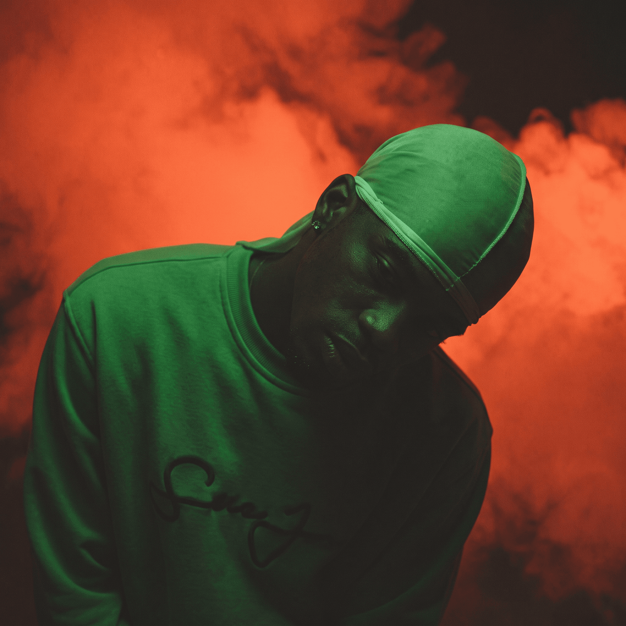 Full-figure portrait of a man in a green sweatshirt and headscarf against a red and smoky background.