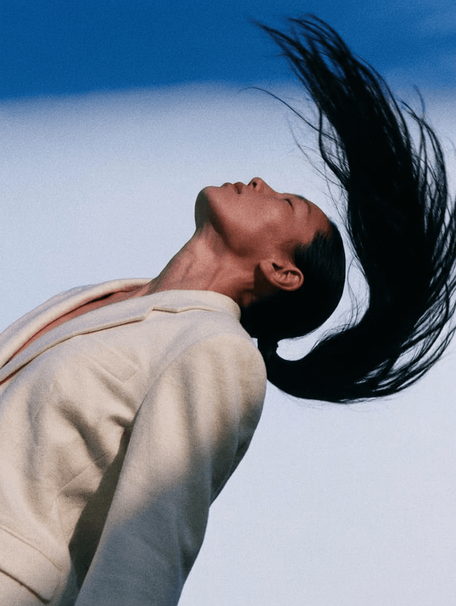 A woman in a full-figure portrait with flowing hair against a blue sky.