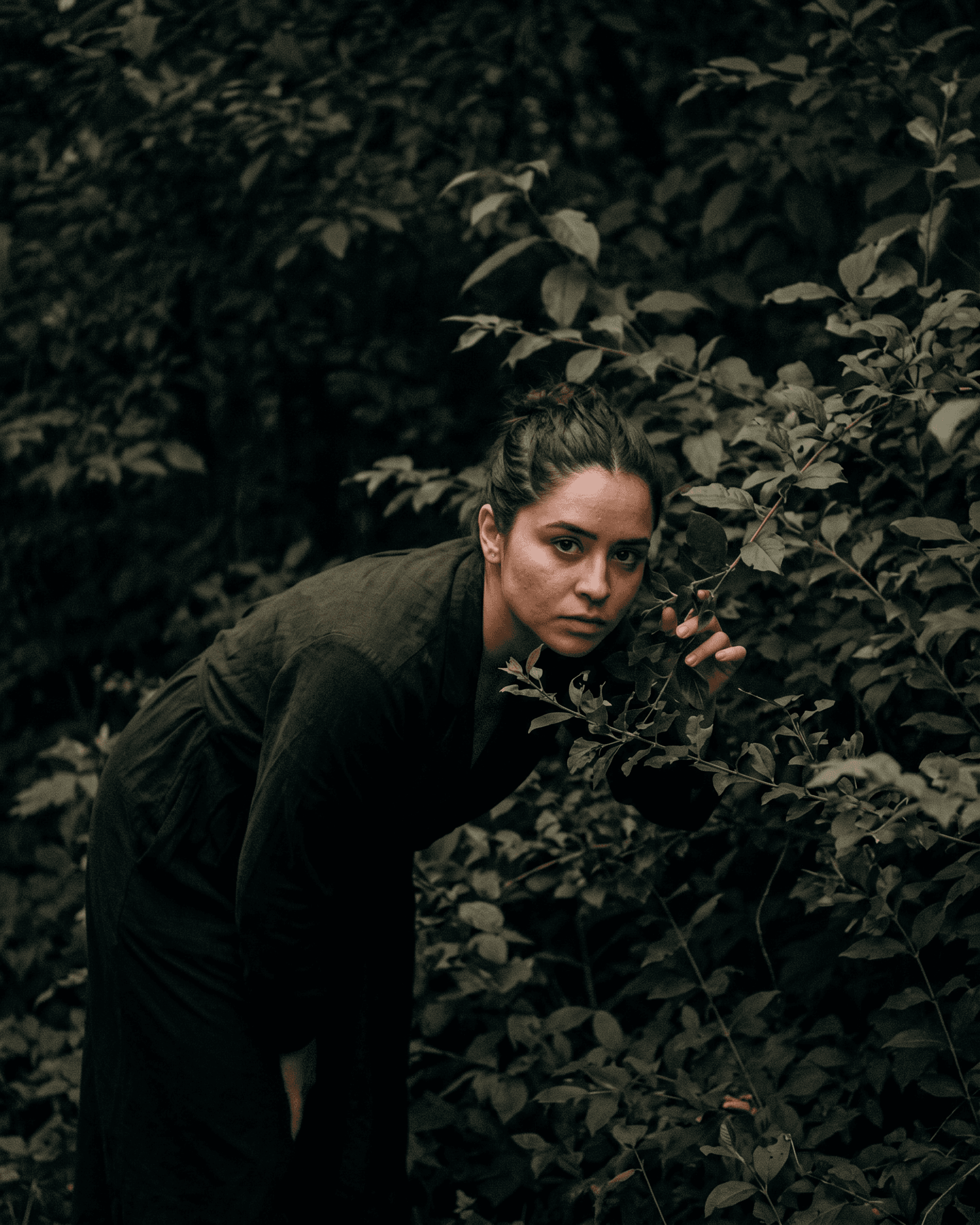 A woman leans towards green foliage in a dark forest.