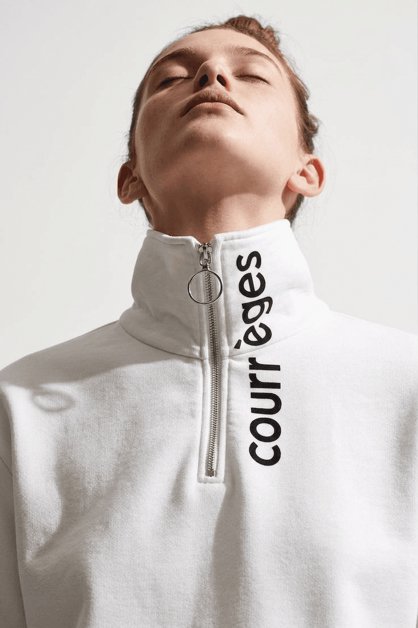 A woman wearing a zippered white sweatshirt with black branding, posing against a clean, minimal background.