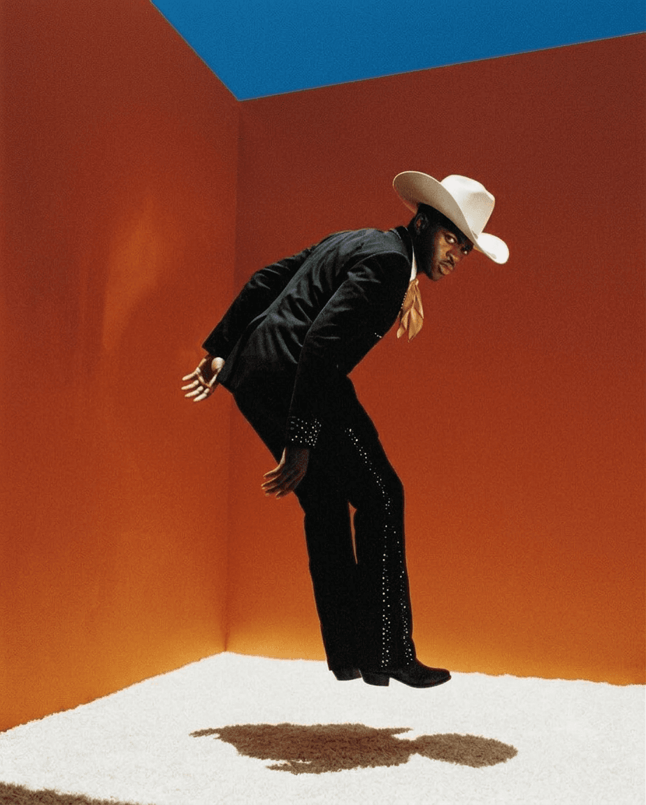 Man in a black suit and cowboy hat jumping against an orange background.