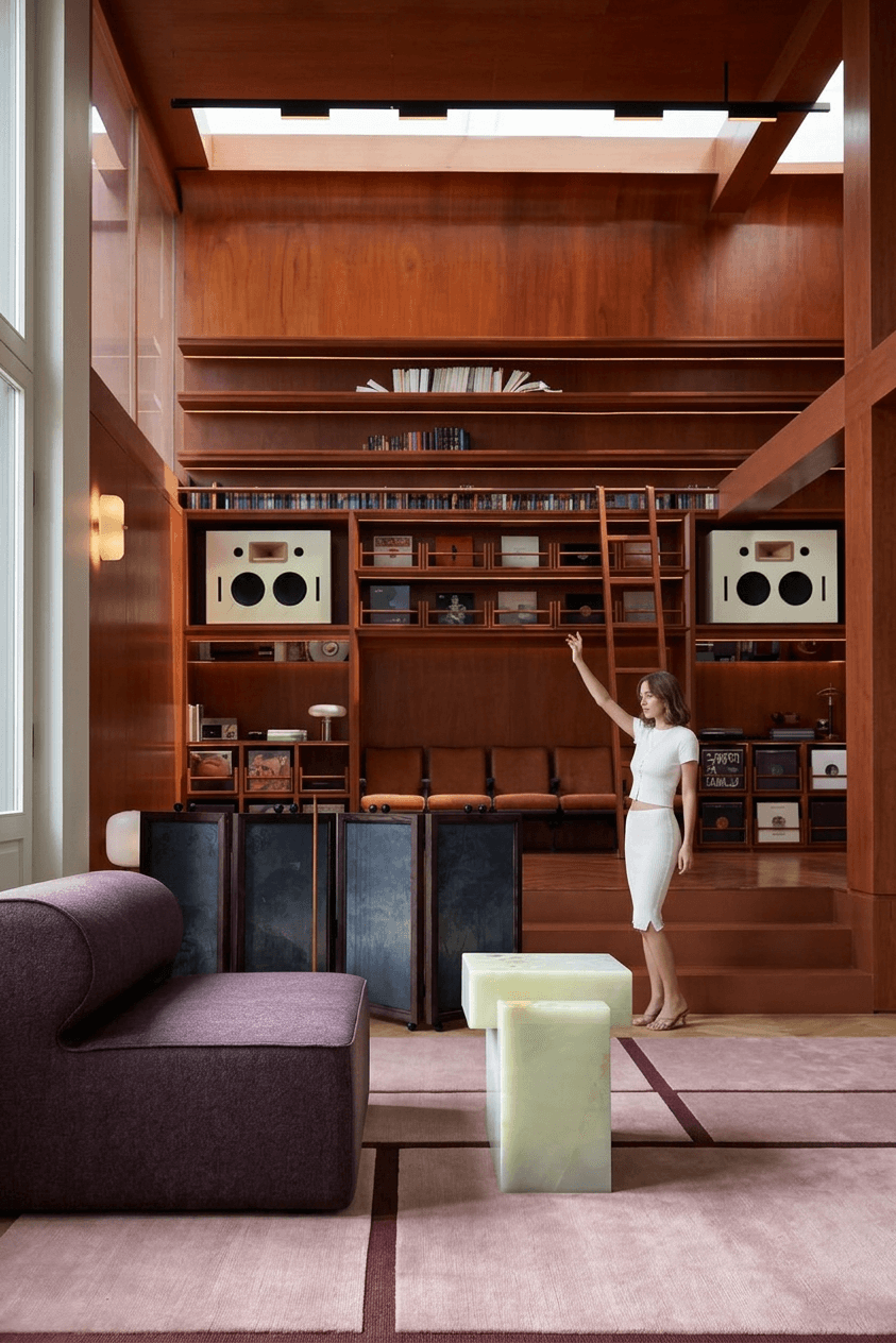 A woman in a fitted white dress stands in a modern interior with wooden walls and stylish furniture.