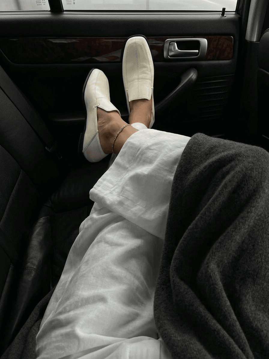 A person in white clothing and cream loafers sitting in a car with a gray blanket.