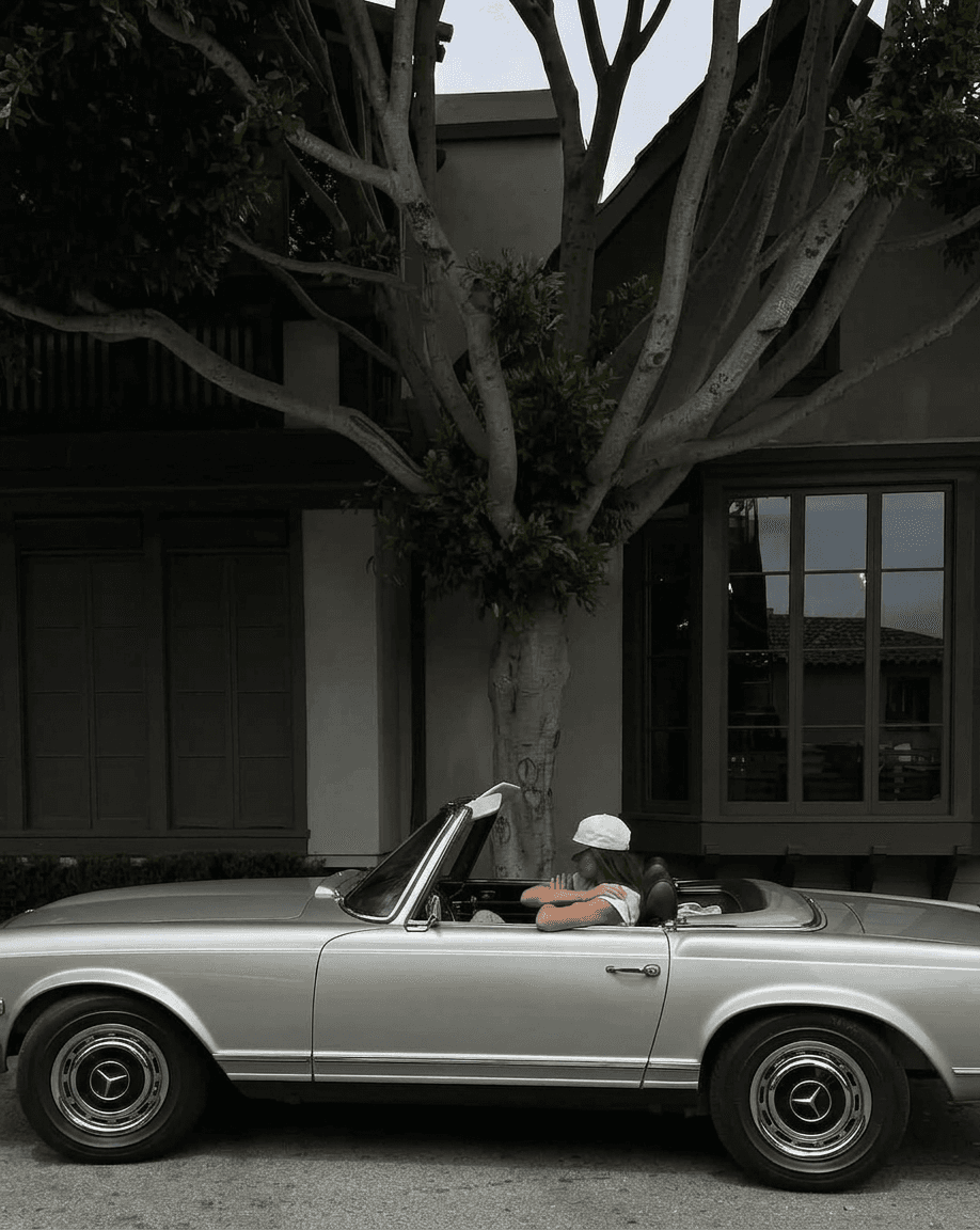 A retro convertible car parked next to a modern house with a person inside.