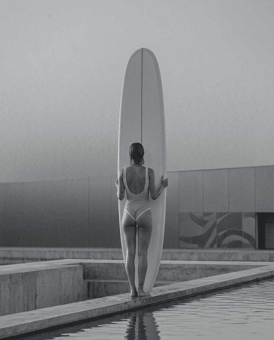 Black-and-white image of a woman in a swimsuit with a surfboard by a calm pool.