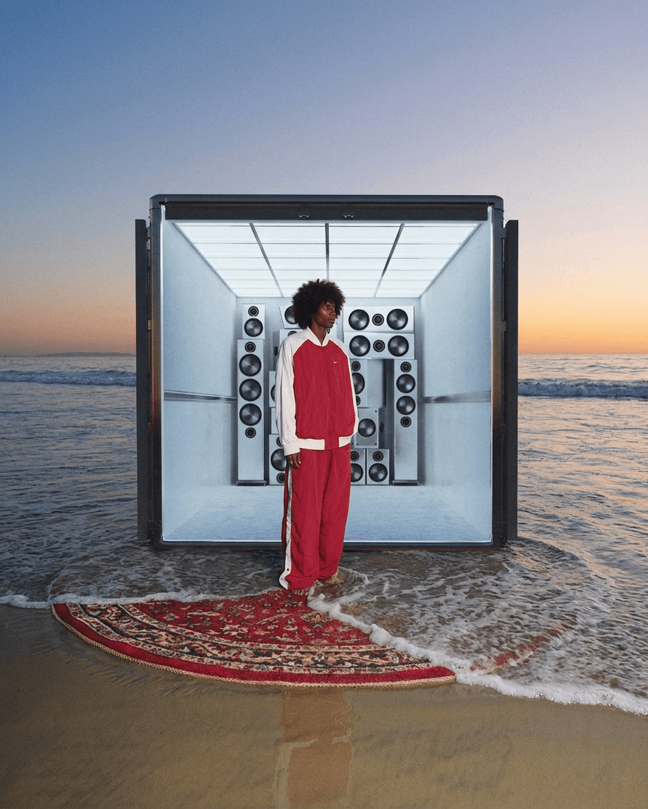 Young person in red tracksuit on rug at shore with sound system.
