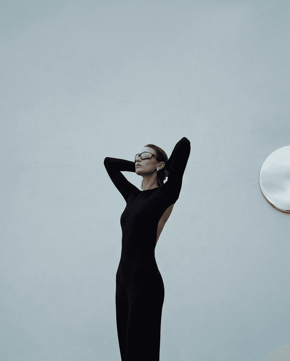 A model in a black jumpsuit and reflective sunglasses poses against a gray wall.