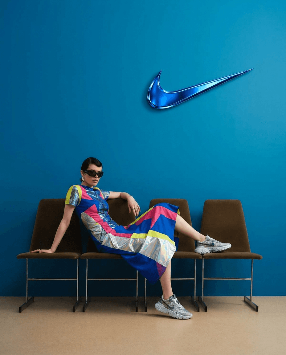 A model wearing a vibrant dress and sunglasses stands against a blue wall with the Nike logo.