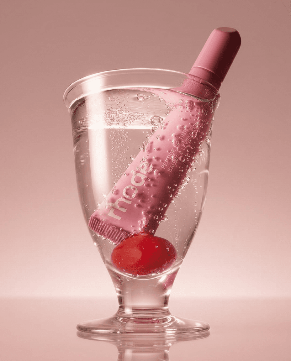 Pink skincare product in a glass of sparkling water with a cherry, on a soft pink background.