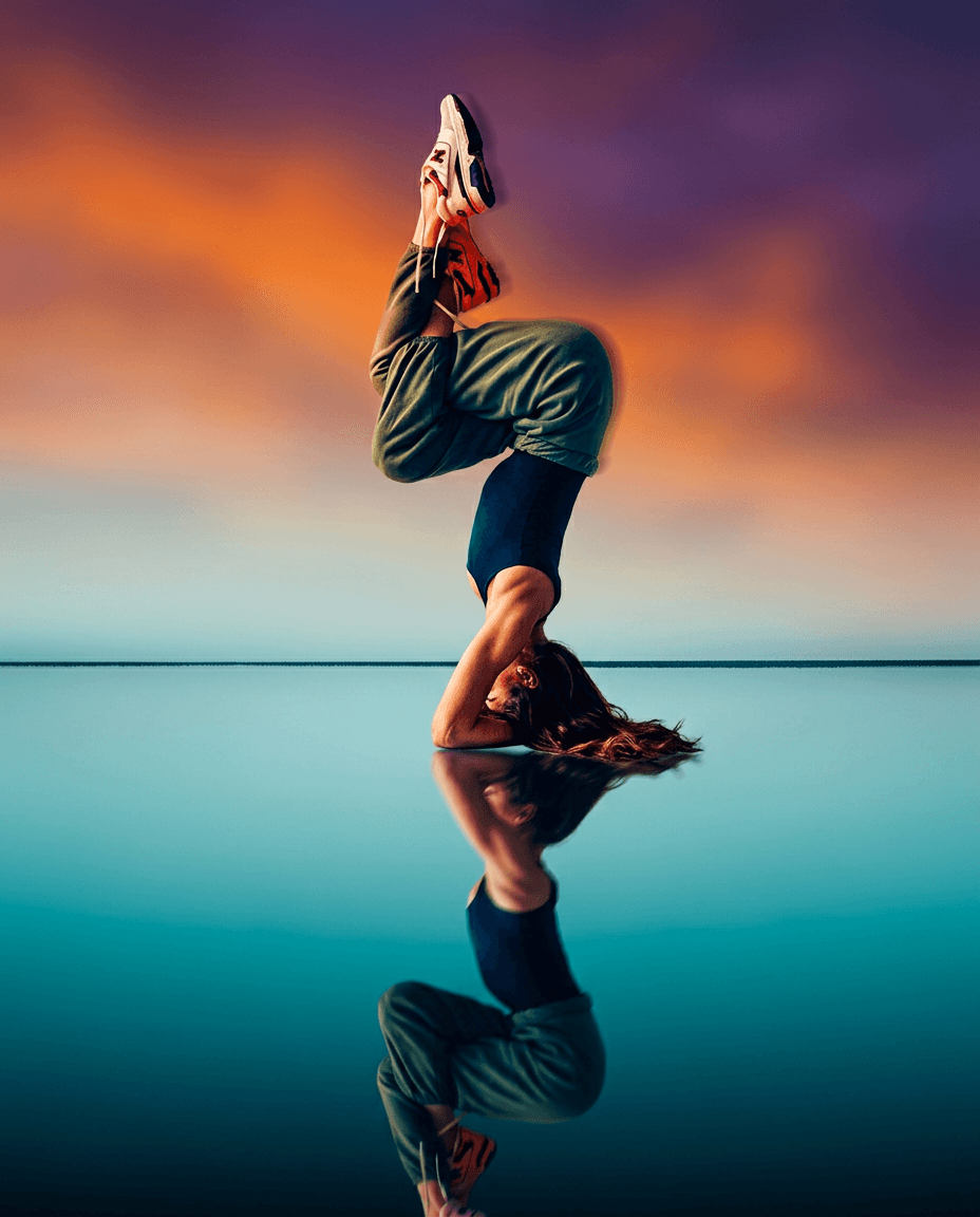 A woman in an upside-down yoga pose reflected on calm water at sunset.