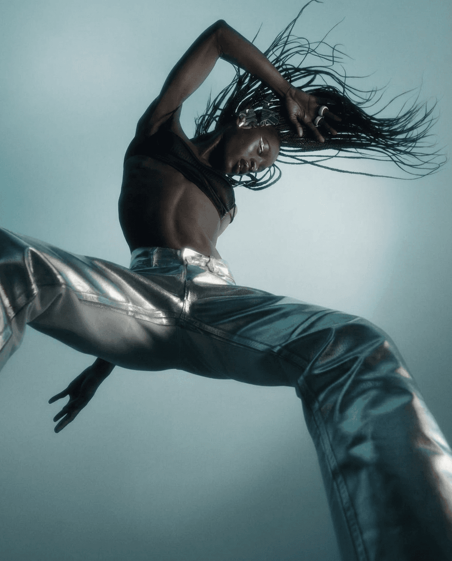 Model wearing metallic silver pants, photographed from below with a dramatic blue and silver background.