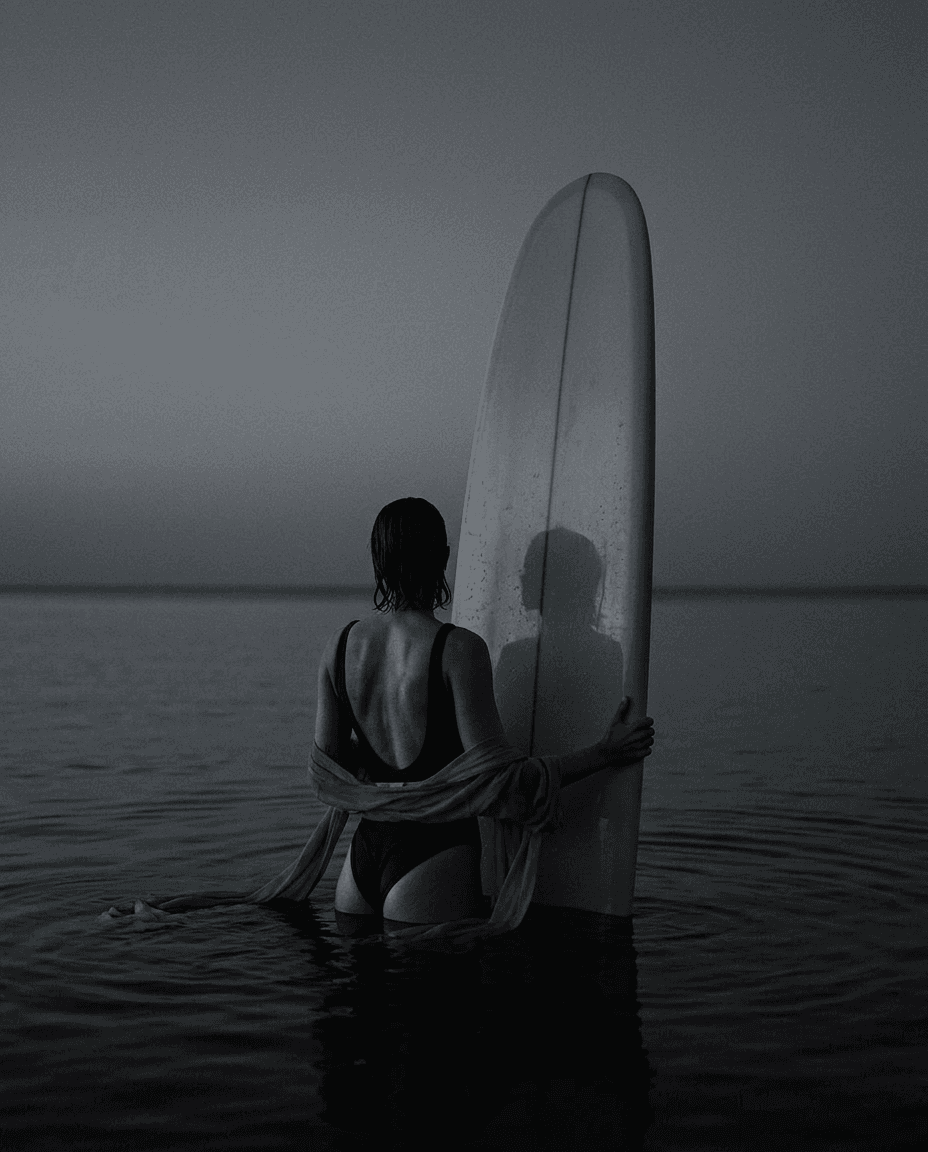 A figure in a swimsuit stands in calm water holding a surfboard, looking towards the horizon.