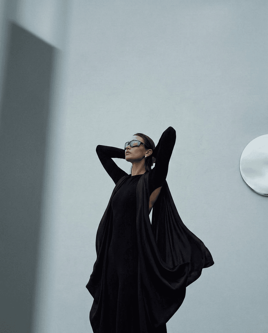 A model wearing a black gown with long sleeves and sunglasses, set against a gray background.