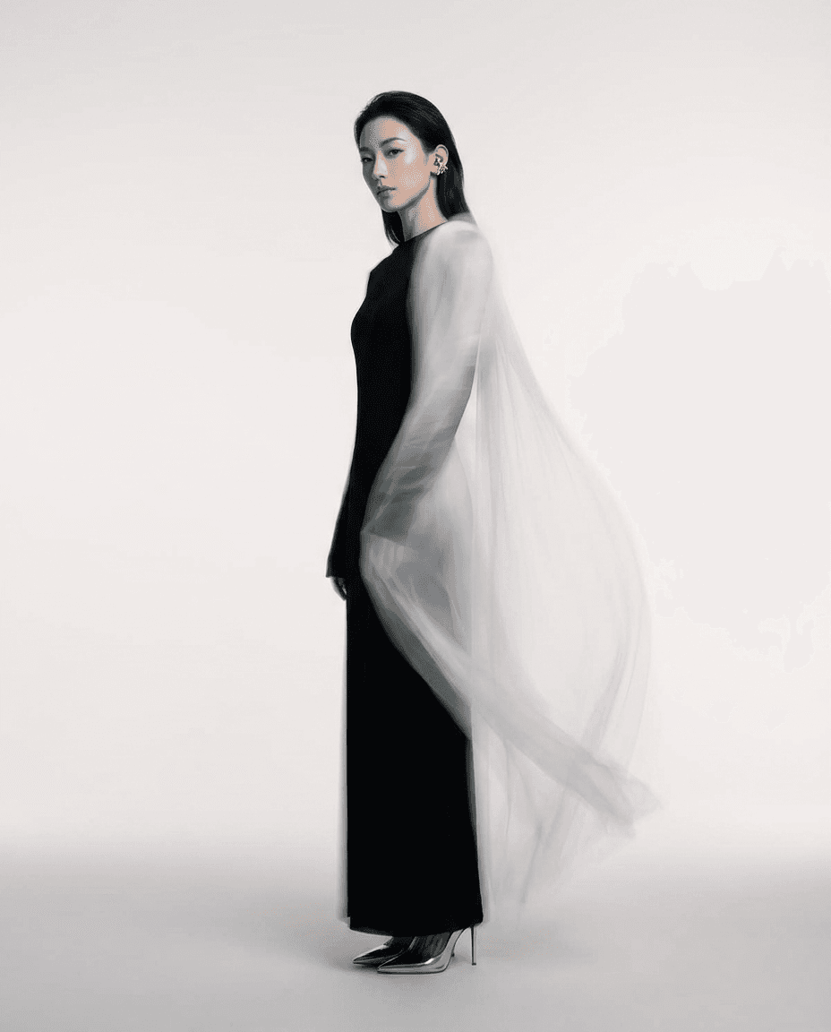 A woman in a black gown and sheer cape stands against a light grey background.