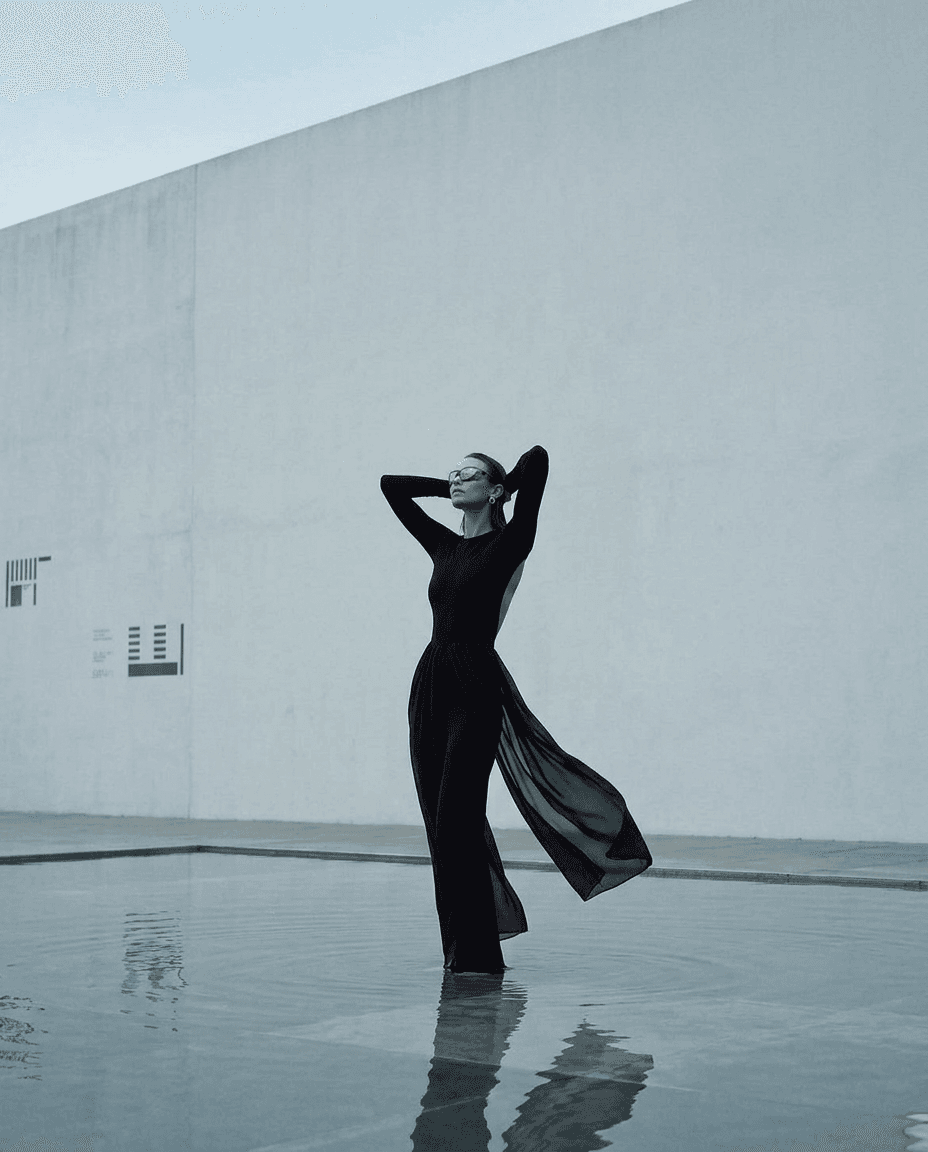 Model in a black outfit standing in shallow water with a white wall behind.