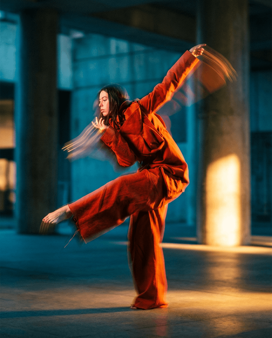 A dancer in an orange outfit poses dynamically in front of concrete columns.