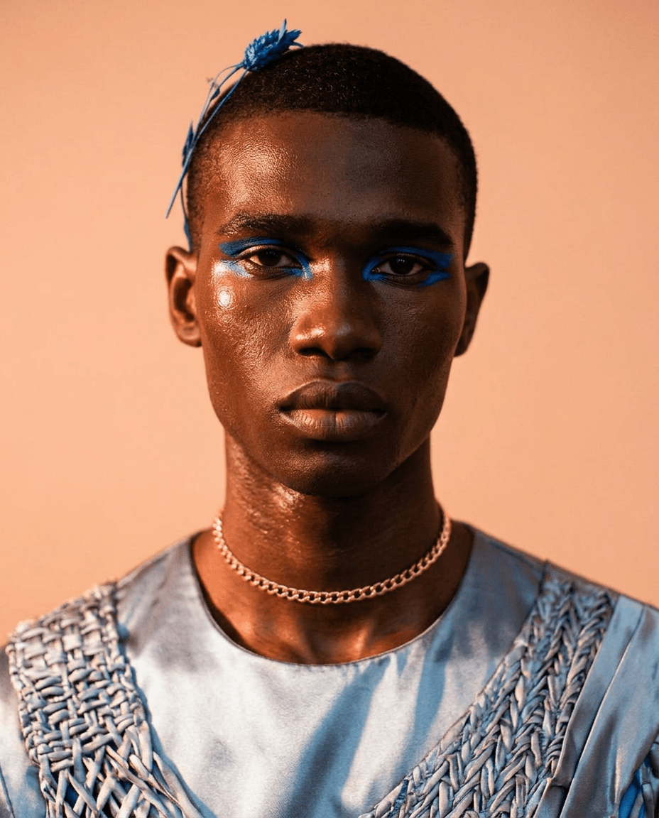 A young man with blue eyeshadow and a hair accessory, set against a soft, warm background.