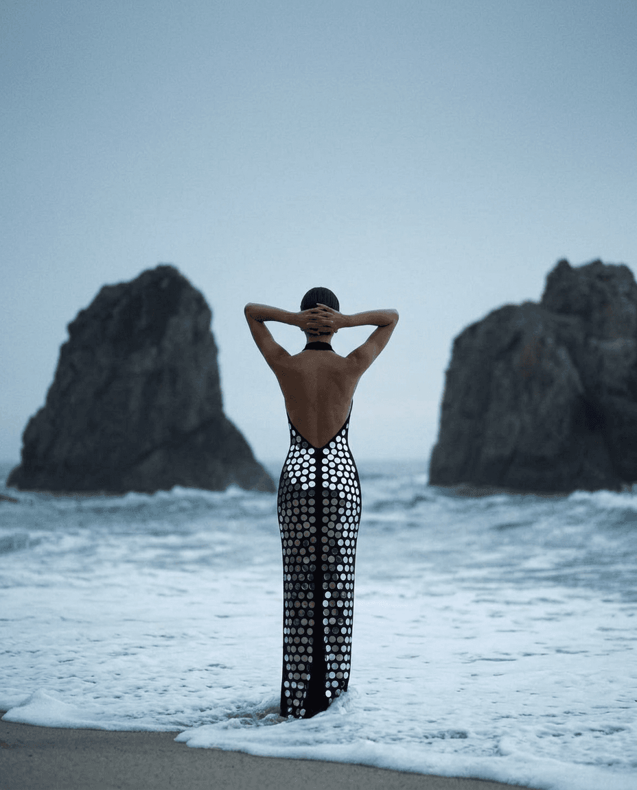 Model in a backless dress with reflective discs by the ocean.
