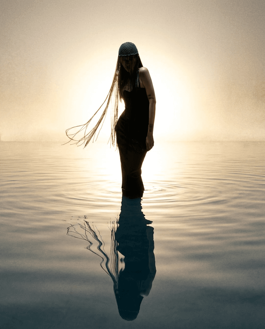 Silhouetted figure in a black dress standing in calm water with a glowing background.