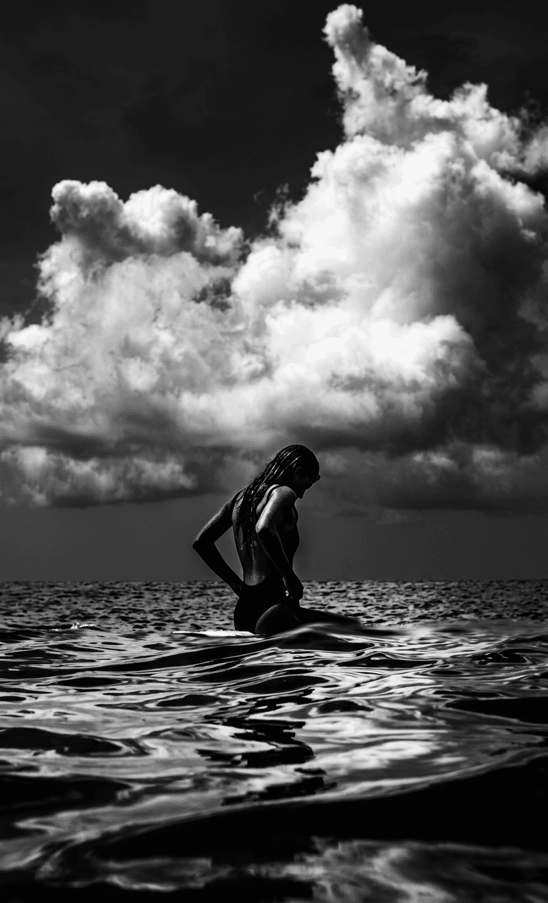 Silhouetted figure wading through calm ocean waters with dramatic clouds above.