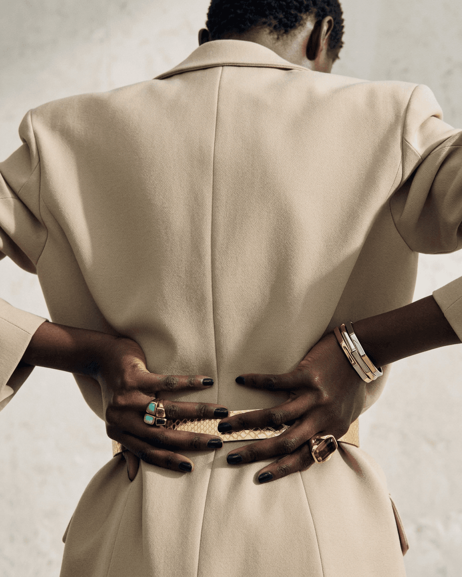 Model wearing a beige tailored coat, displaying statement rings and bracelets.