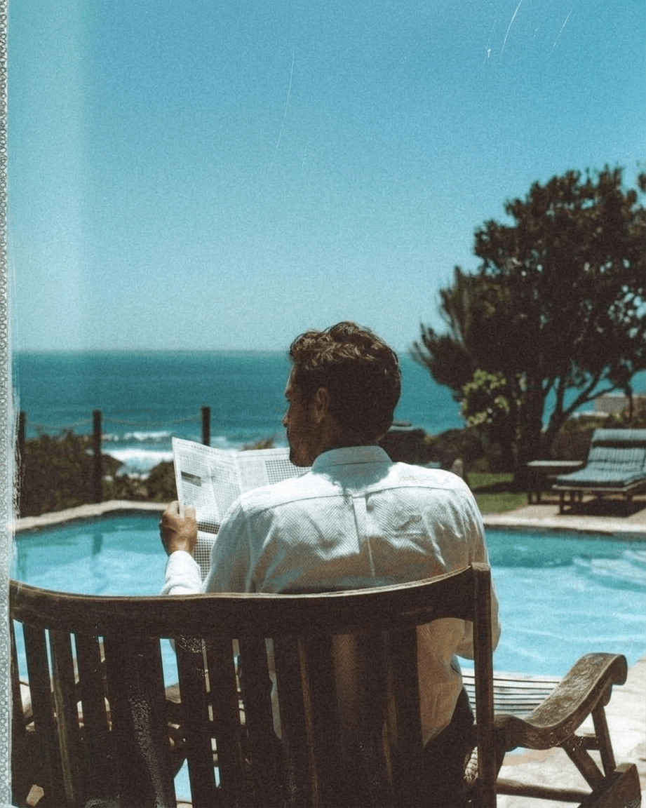 Man in a white shirt sitting by a pool, reading a newspaper with ocean view.