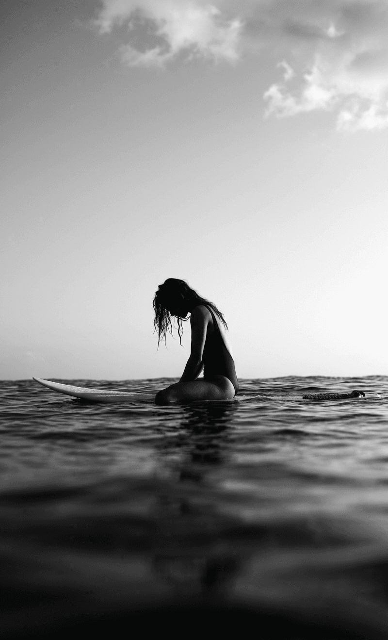 A lone surfer sitting on a board in the water under a cloudy sky.