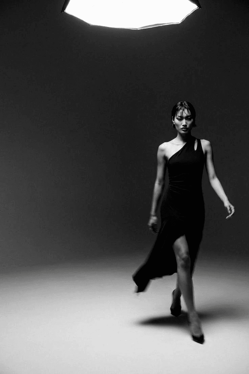 A confident woman in a one-shoulder dress walking under a spotlight.