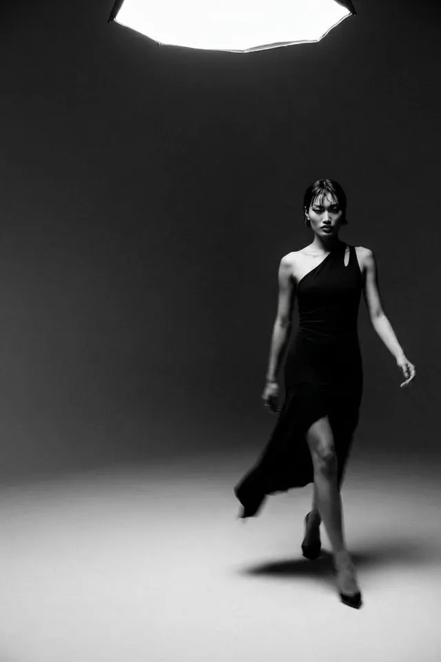 A confident woman in a one-shoulder dress walking under a spotlight.