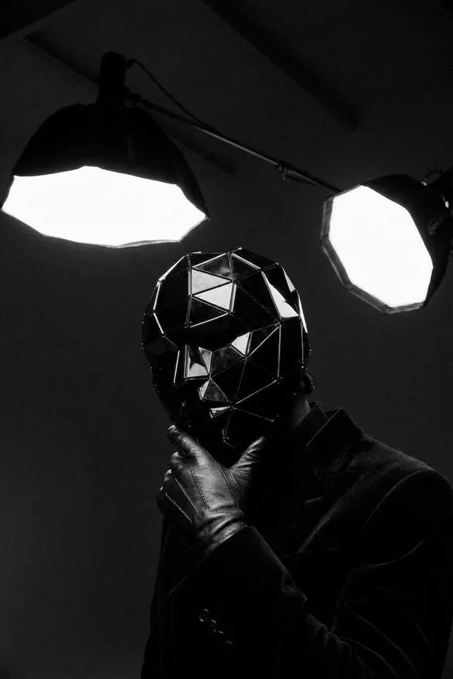 A figure wearing a geometric black mask poses under bright spotlights.