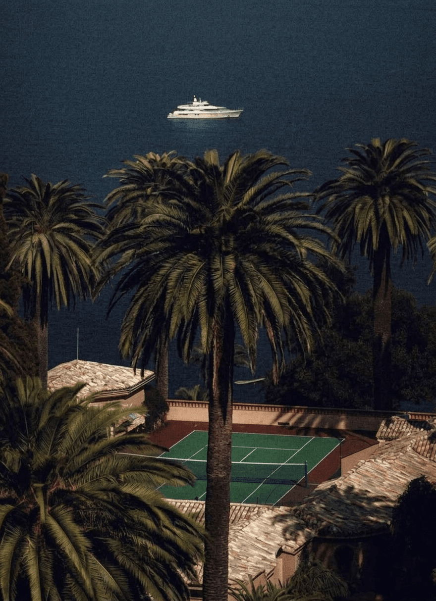 Aerial view of a coastal area with palm trees, a tennis court, and a yacht in calm waters.