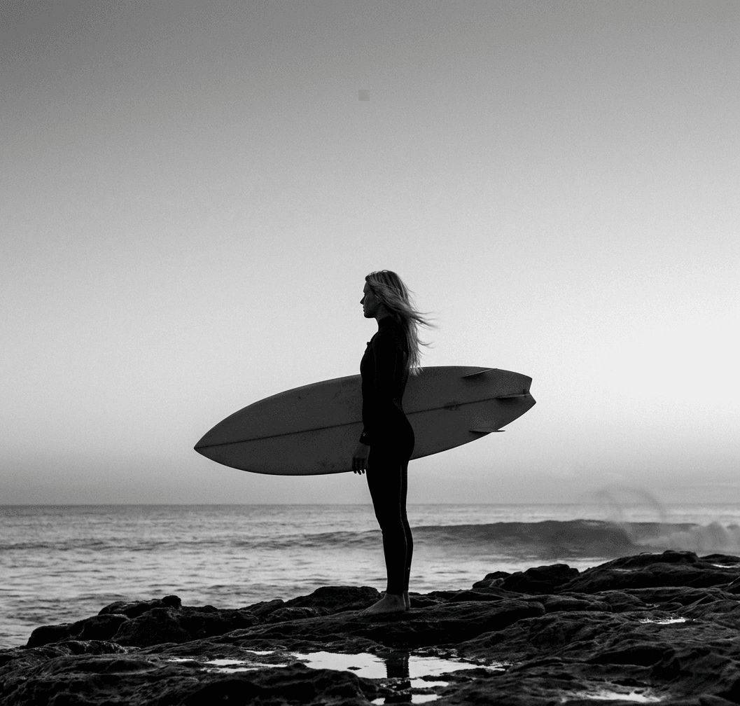 Black and white silhouette of a woman standing on rocks with a surfboard by the ocean.