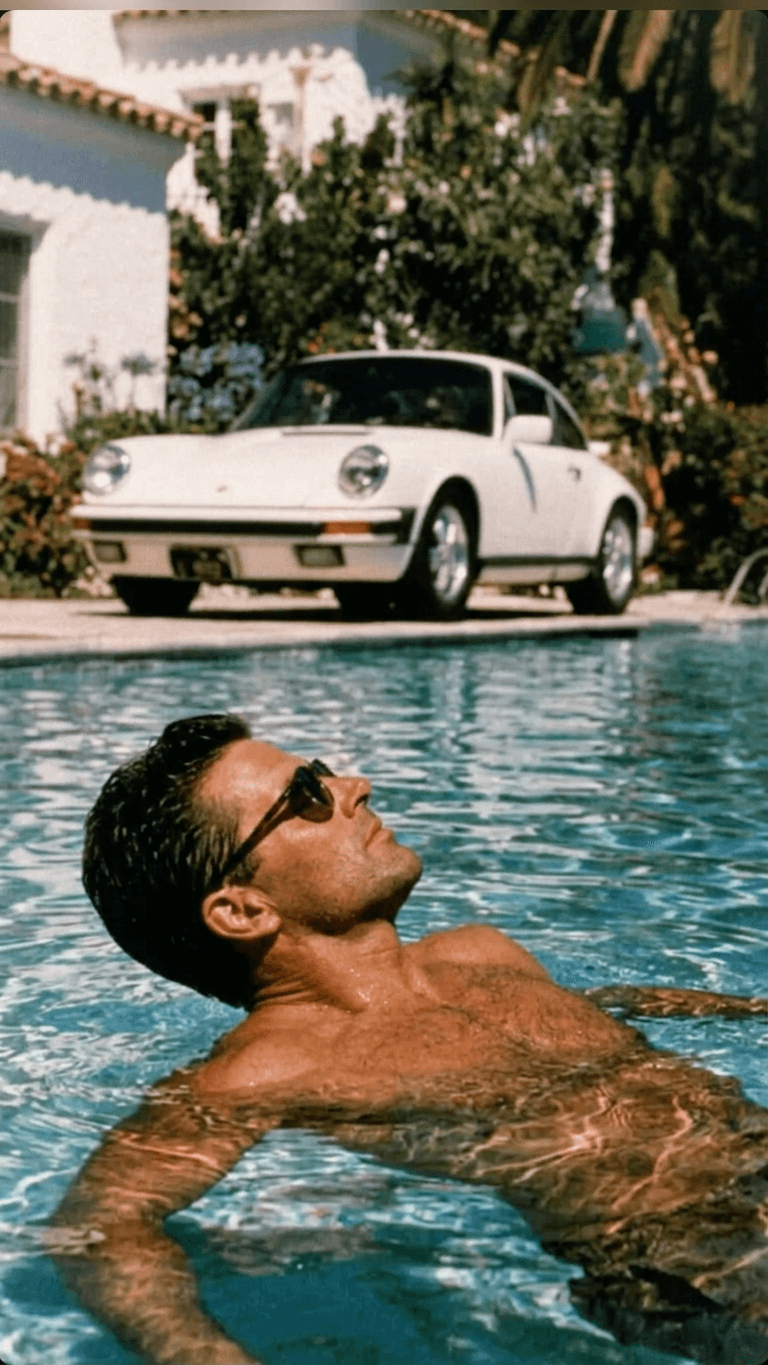 A man wearing sunglasses floats in a blue pool near a white Porsche and greenery.