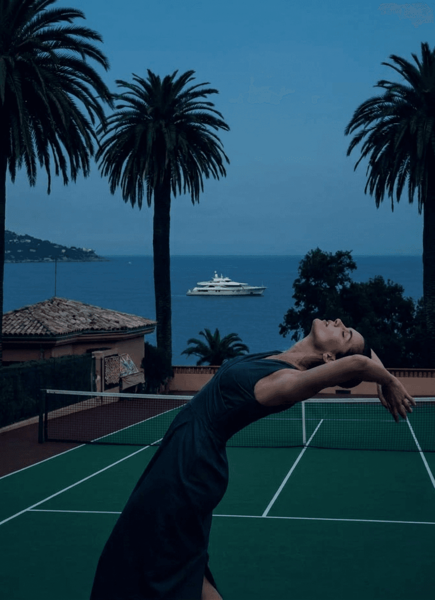 A woman in a dark dress poses on a tennis court with palm trees and the ocean in the background.