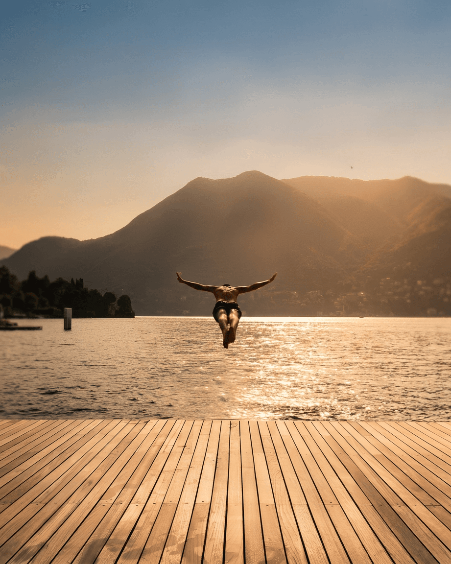 Silhouette of a person diving from a dock into a calm lake with mountains behind.