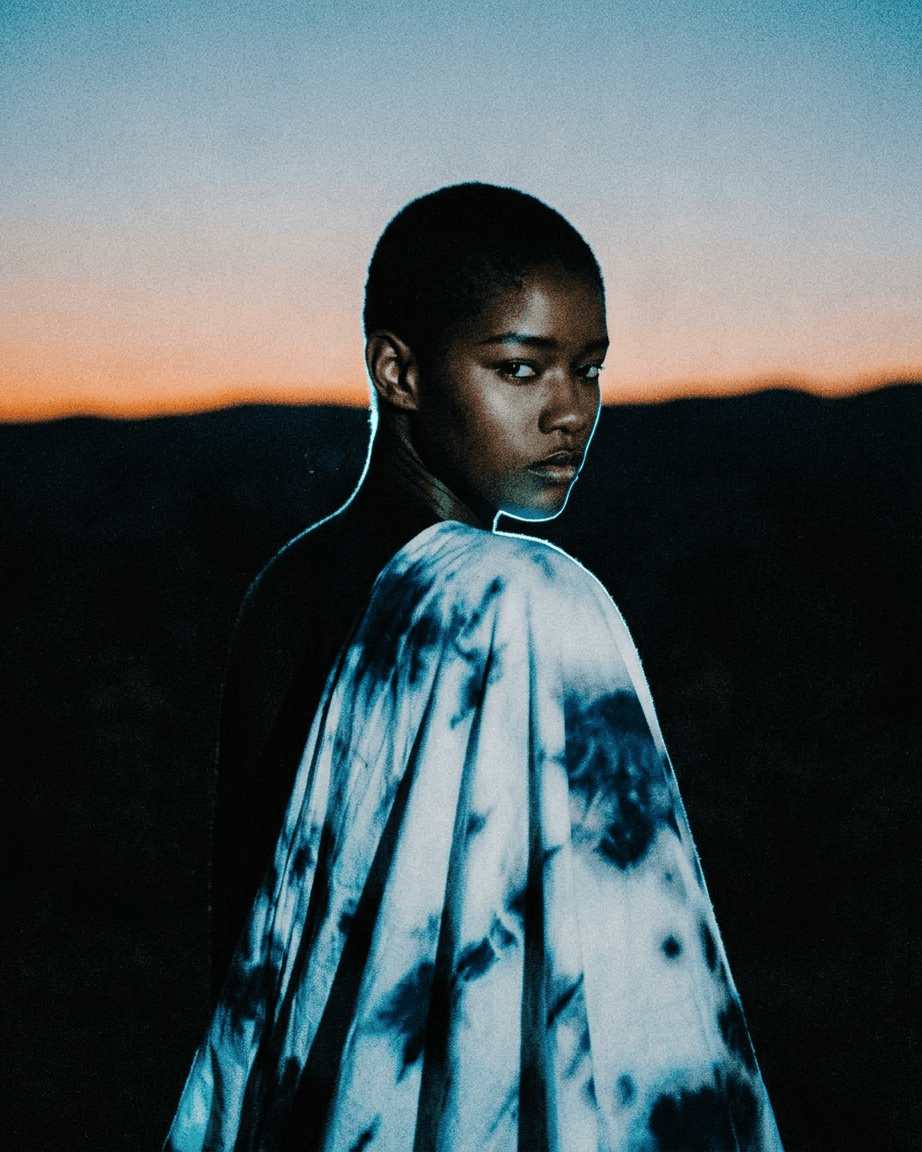 Portrait of a model with a short haircut draped in tie-dye fabric against a twilight background.