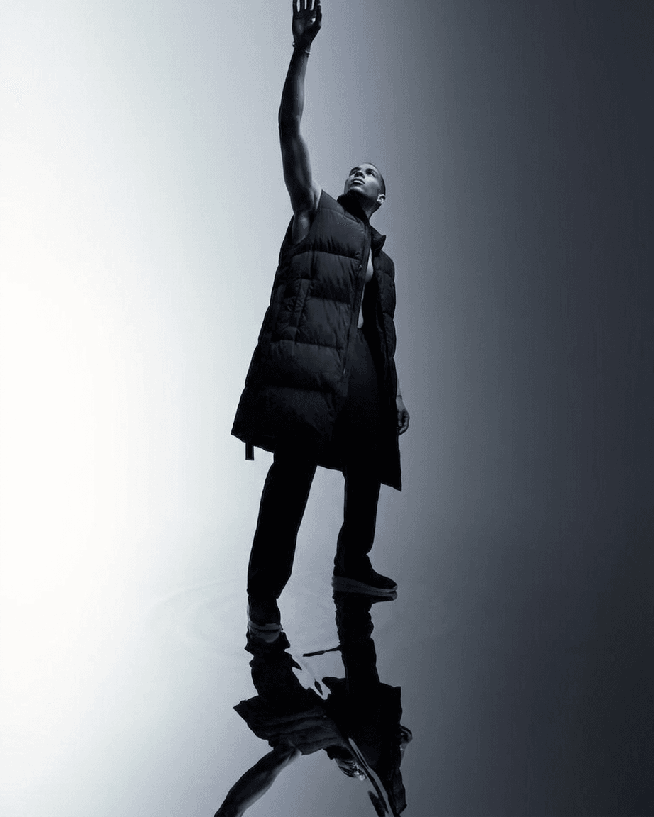 Model wearing a sleeveless black puffer coat reaching upward against a reflective background.