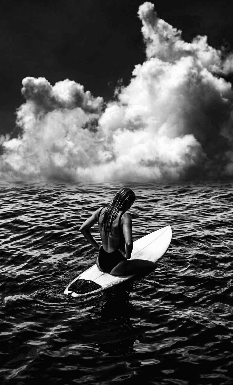 A surfer sits on a surfboard in dark waters with dramatic clouds above.