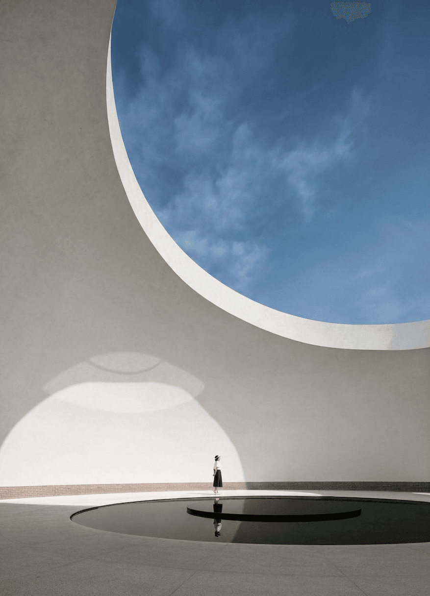 A figure by a circular pool beneath a large circular opening in a white architectural structure.